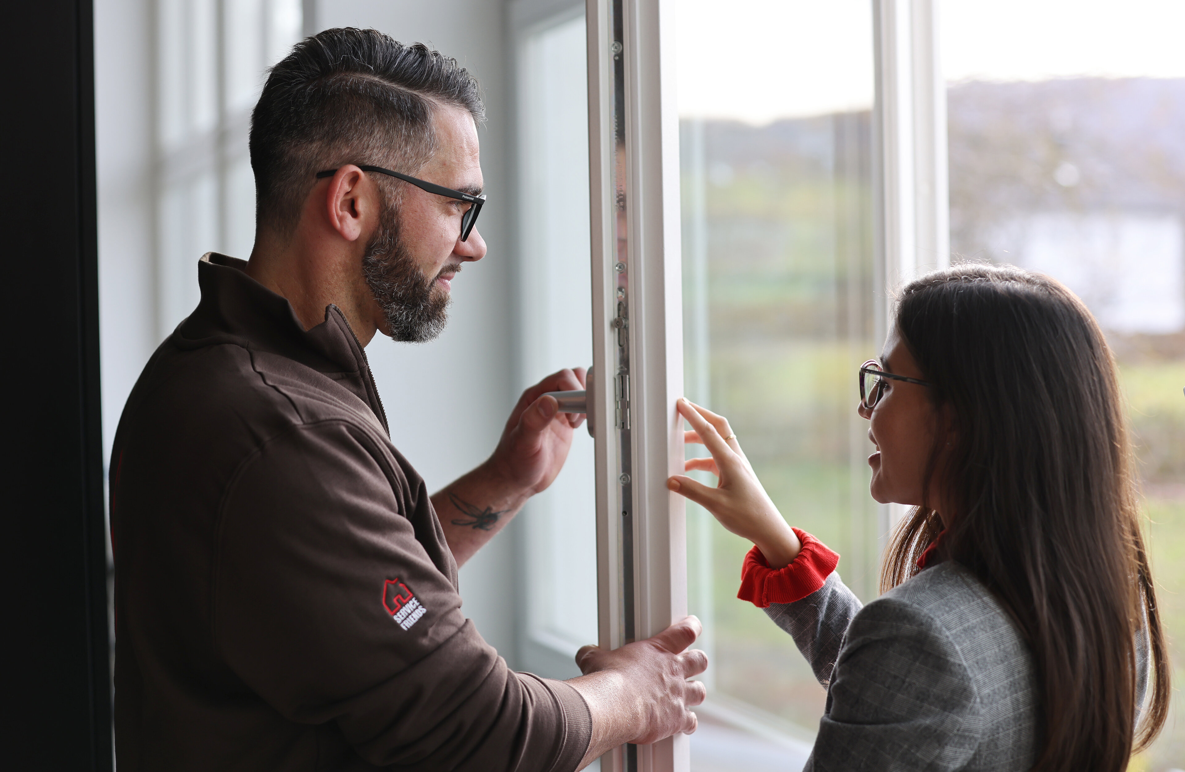 Ein Mann und eine Frau die sich angucken. Die Frau berührt den Rahmen des Fensterflügels und der Mann hält den Fensterflügel und den Fenstergriff fest. 